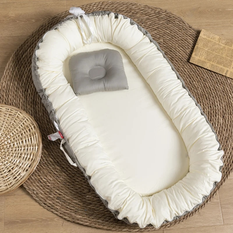 A gray and white padded cotton baby lounger with a pillow on a flat surface, surrounded by a woven basket.
