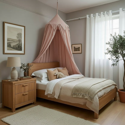 A styled bedroom featuring a light wood bed with a dusty pink fabric canopy, dressed in neutral beige and white bedding. The room has gray walls, a wooden nightstand with a lamp, a white rug, and a potted olive tree by the window.