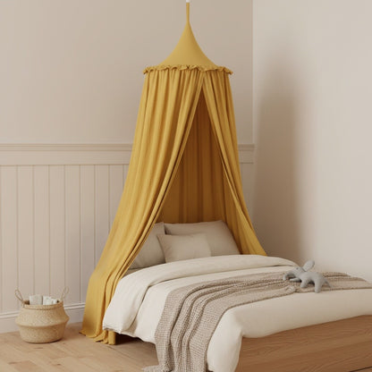 A mustard yellow fabric canopy bed in a styled children's bedroom. The bed is dressed in white bedding with a textured beige throw blanket, set against a cream wall with white wainscoting.
