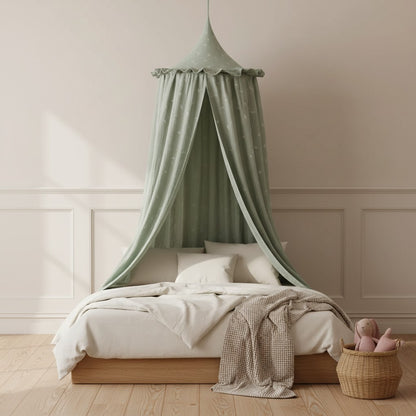 A light sage green baby canopy with white star prints hangs over a wooden bed frame in a softly lit nursery. The bed is dressed in white bedding with a textured beige throw blanket, and a small woven basket with pink plush toys sits on the floor nearby.