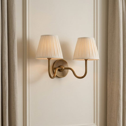 A collage showing a two-light wall sconce in multiple room settings. The fixture has a warm brass or bronze curved arm and backplate with two pleated off-white fabric shades.