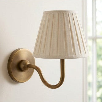 A collage showing a two-light wall sconce in multiple room settings. The fixture has a warm brass or bronze curved arm and backplate with two pleated off-white fabric shades.