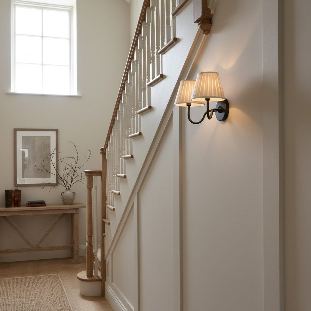 A two-light wall sconce with beige pleated shades and a black metal mount, shown illuminated on a cream-colored interior wall next to a wooden staircase.