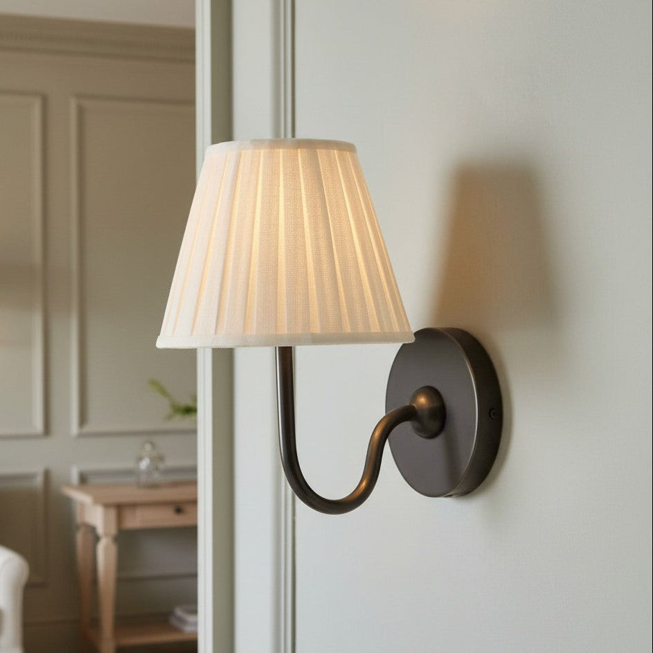 A wall-mounted sconce with a pleated off-white fabric lampshade and a dark bronze curved metal arm, shown lit on a white paneled wall in a softly styled interior.
