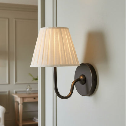 A wall-mounted sconce with a pleated off-white fabric lampshade and a dark bronze curved metal arm, shown lit on a white paneled wall in a softly styled interior.