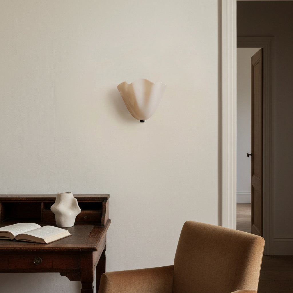 A modern wall sconce with a fluted, organic-shaped cream-colored glass shade mounted on a warm off-white wall in a styled interior room with a wooden desk, chair, and vase.