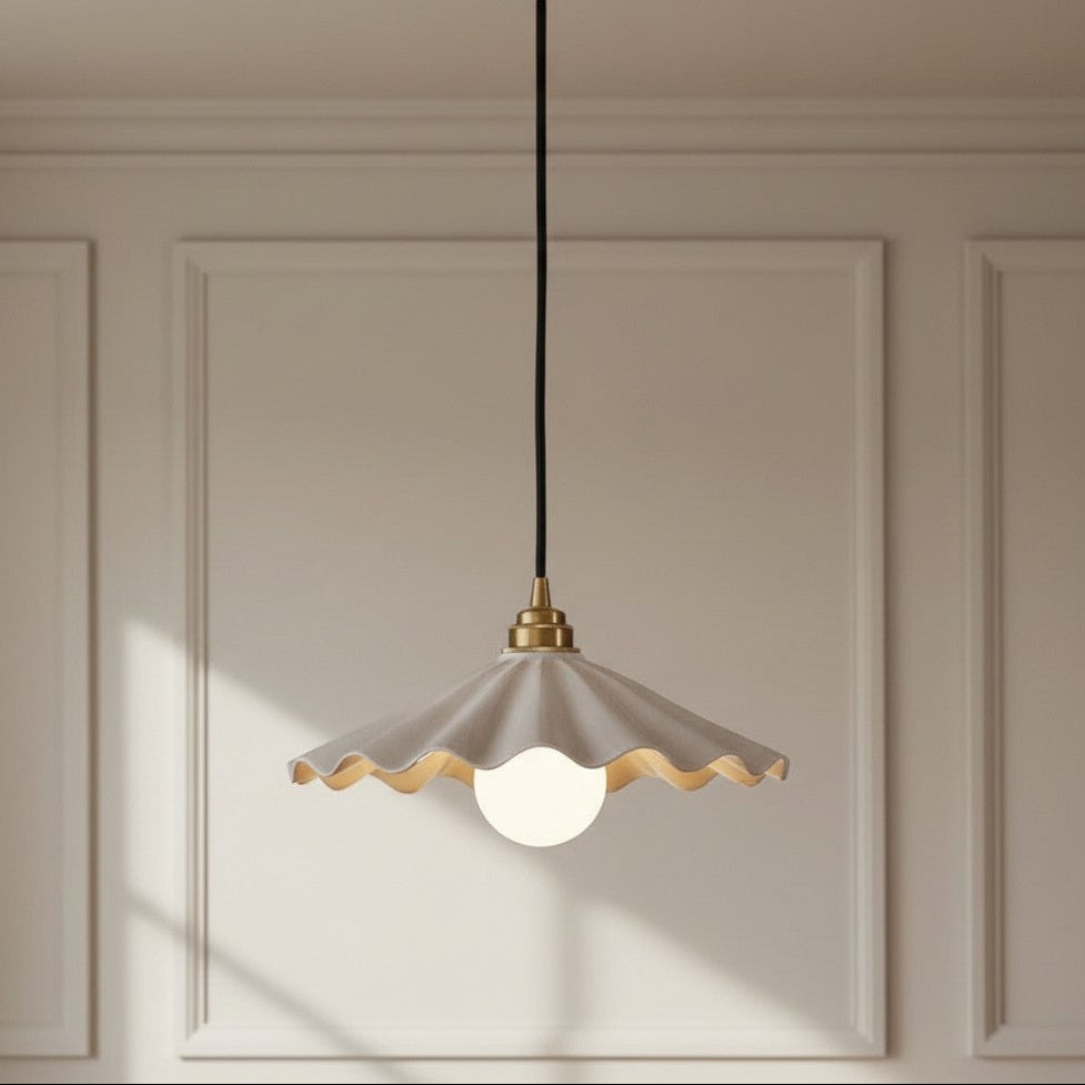A modern pendant light with a scalloped off-white shade, gold-toned brass hardware, and a black cord, hanging against a beige paneled wall.