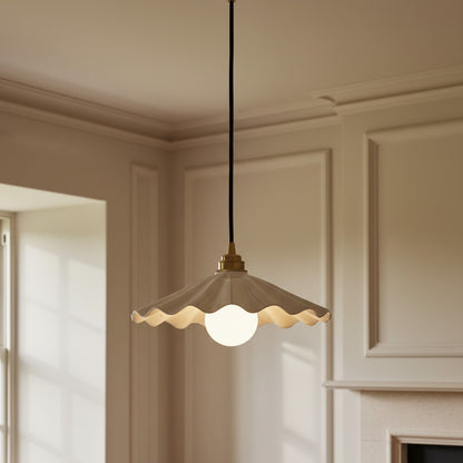 A hanging pendant light with a scalloped cream glass shade, a white globe bulb, and a brass fixture, suspended by a black cord in a cream-colored room with molding and a fireplace.