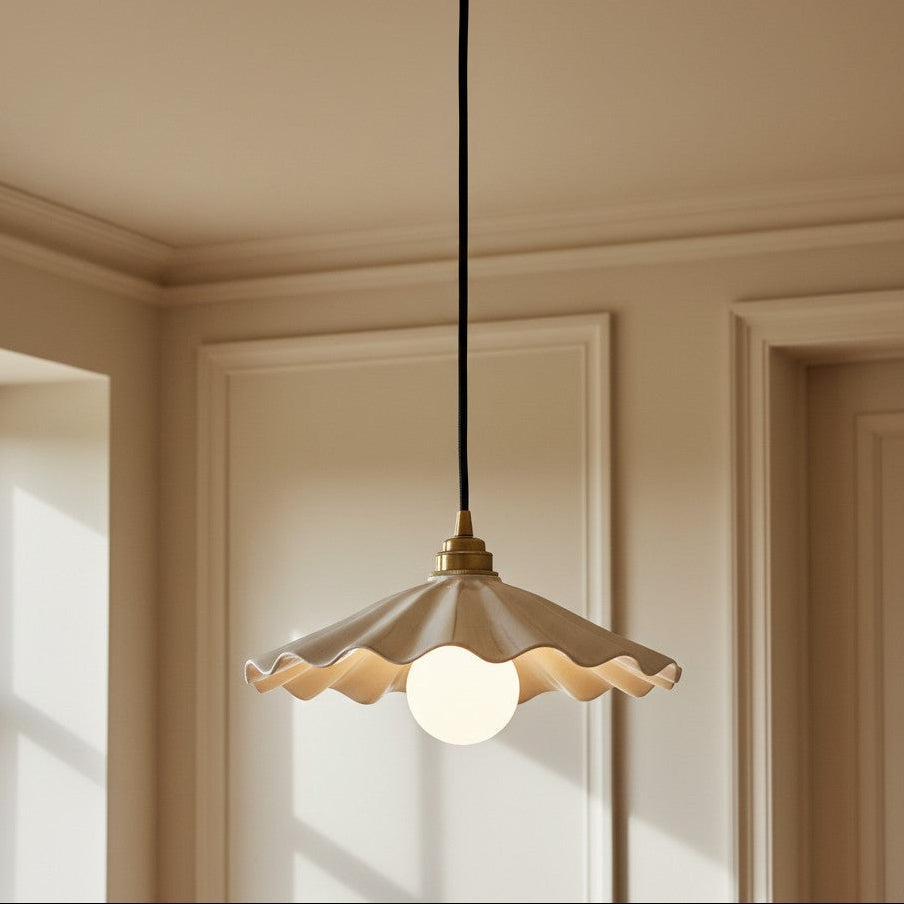 A modern pendant light with a wavy scalloped beige shade, a black cord, and a brass ceiling canopy, shown hanging in a warm beige room.