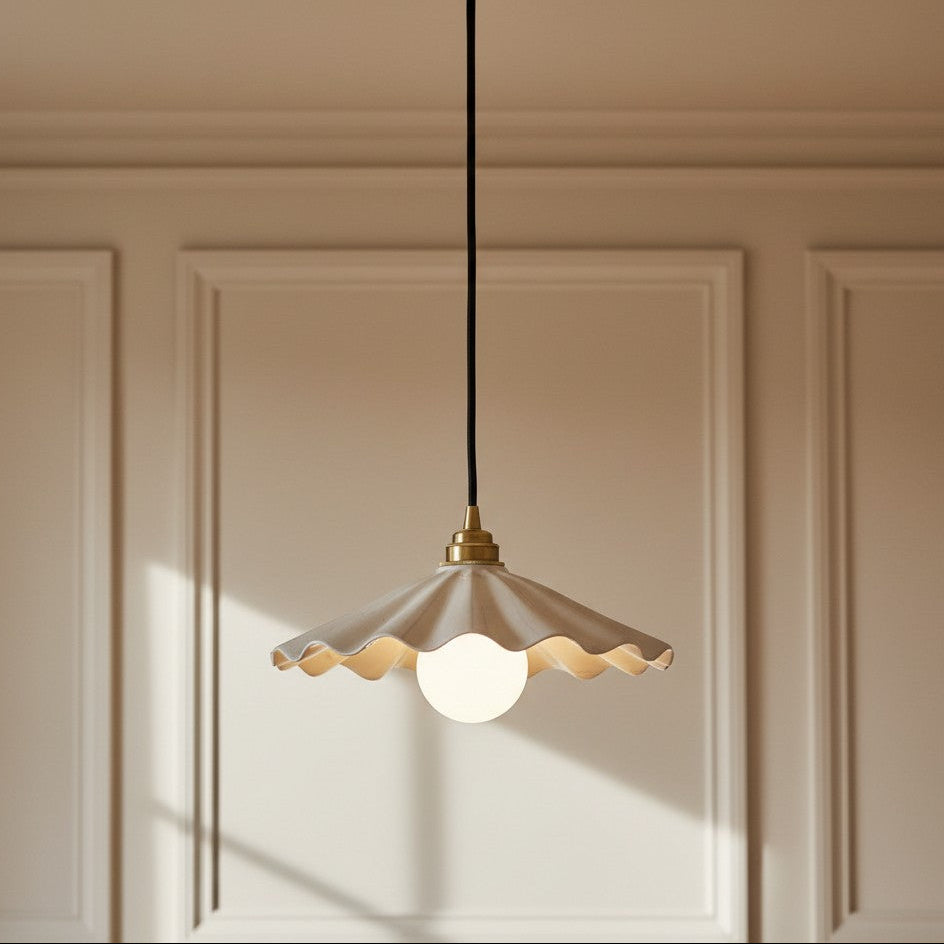 A modern pendant light with a scalloped white glass shade, brass metal hardware, and a black cord, hanging against a beige paneled wall.