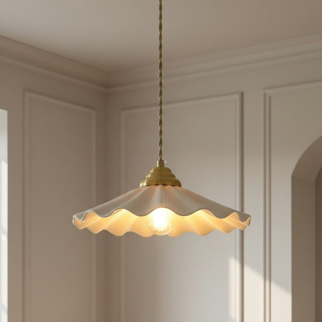 A hanging pendant light with a scalloped cream glass shade and a brass-toned metal top and cord, shown lit in a softly styled interior room with paneled walls.