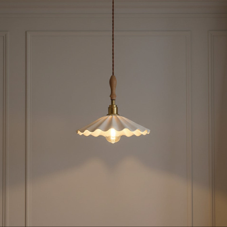A hanging pendant light with a scalloped white glass shade, brass metal hardware, and a light wood finial, suspended against a beige paneled wall.
