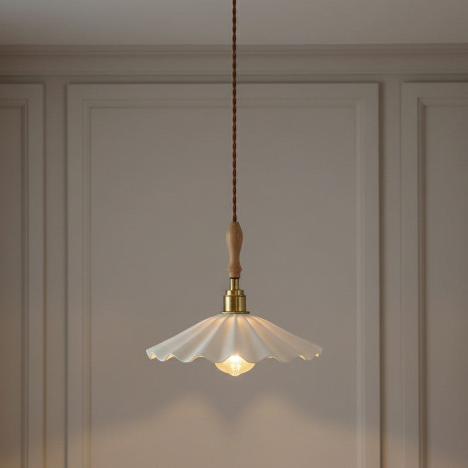 A hanging pendant light with a white scalloped glass shade, a light wood finial, and a brass socket, suspended by a braided brown cord against a beige paneled wall.