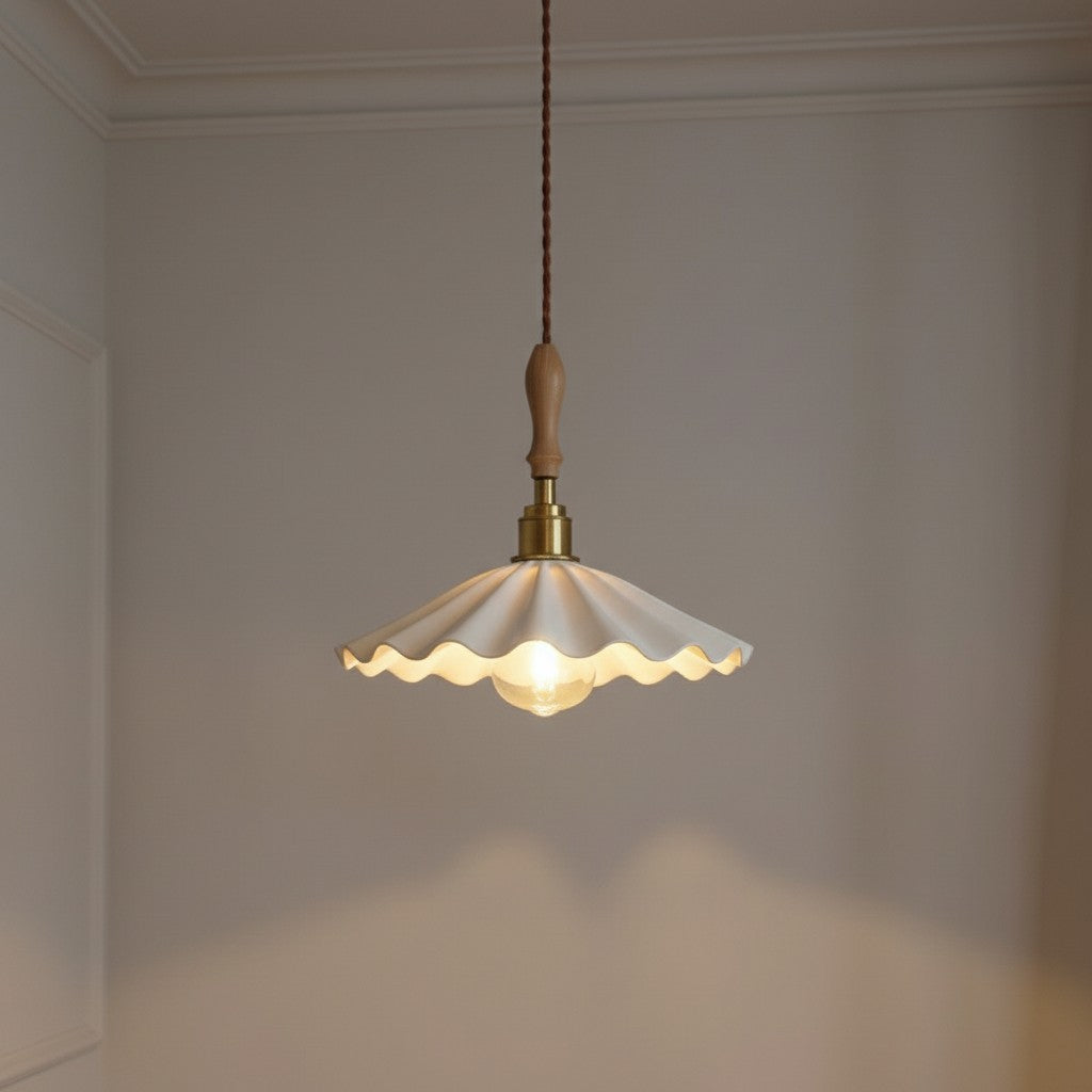 A vintage-style pendant light with a scalloped cream glass shade, brass metal fixture, and a light wood finial, hanging against a neutral beige wall.