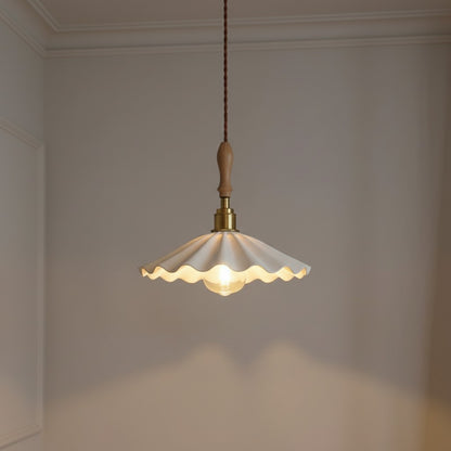 A vintage-style pendant light with a scalloped cream glass shade, brass metal fixture, and a light wood finial, hanging against a neutral beige wall.