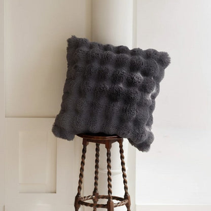 Dark gray fluffy pillow on a wooden stool in a room with a desk and chair.